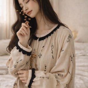 May include: Beige pajama set with black floral print. The long-sleeved top has a black lace trim on the collar and cuffs. The fabric appears soft and comfortable, suitable for sleepwear. The set is button-down.