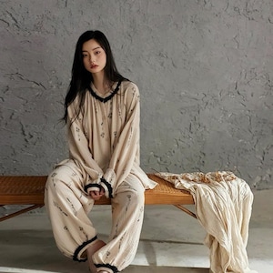 May include: A beige pajama set with a floral pattern and black trim. The long-sleeved top and pants are worn by a person sitting on a wooden bench. A matching beige blanket is draped on the bench.