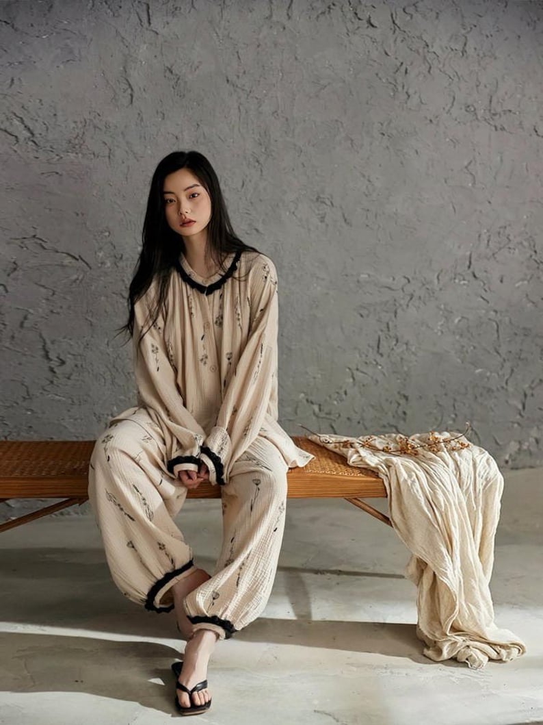 May include: A beige pajama set with a floral pattern and black trim. The long-sleeved top and pants are worn by a person sitting on a wooden bench. A matching beige blanket is draped on the bench.