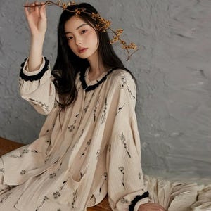 May include: Beige pajama set with black floral print. The long-sleeved top has a black ruffled collar and cuffs. The matching pants have a relaxed fit. The model is holding a branch with small yellow flowers.