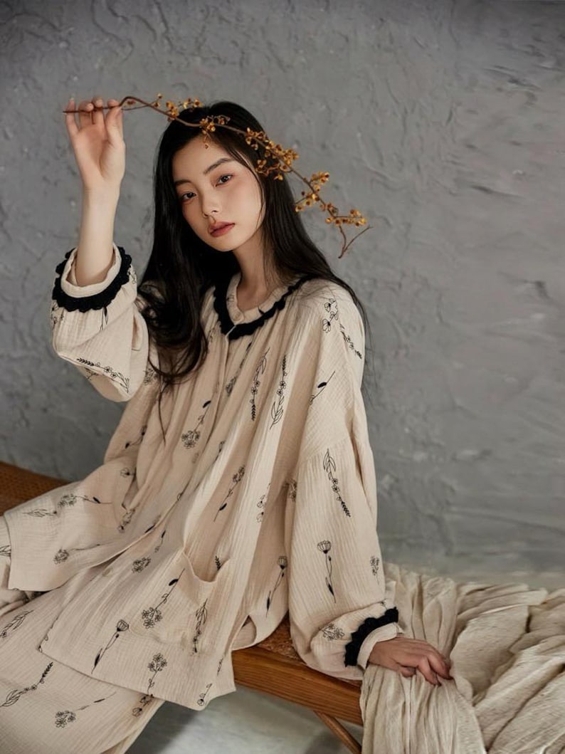 May include: Beige pajama set with black floral print. The long-sleeved top has a black ruffled collar and cuffs. The matching pants have a relaxed fit. The model is holding a branch with small yellow flowers.