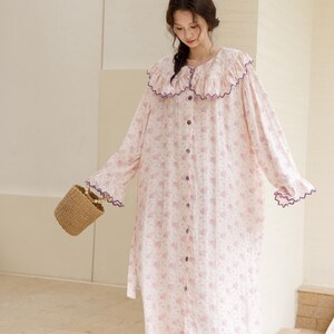 May include: A long, button-down nightgown with a floral pattern in shades of pink and white. The nightgown features a ruffled collar and cuffs trimmed with purple. A small woven basket and brown sandals complete the look.