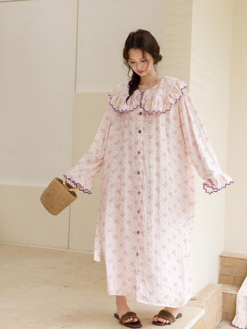 May include: A long, button-down nightgown with a floral pattern in shades of pink and white. The nightgown features a ruffled collar and cuffs trimmed with purple. A small woven basket and brown sandals complete the look.
