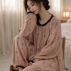 May include: A light pink pajama set with a floral pattern. The long-sleeved top and pants have black trim and buttons. The model is wearing the pajama set while sitting on a wooden chair.