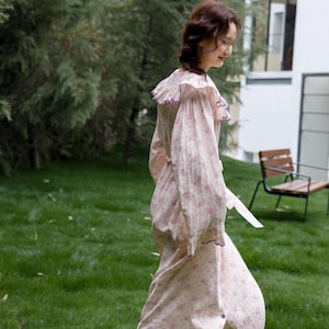 May include: A light pink floral print dress with a ruffled collar and long sleeves. The dress has a button-down front and a matching belt. The dress is worn outdoors on a grassy lawn.