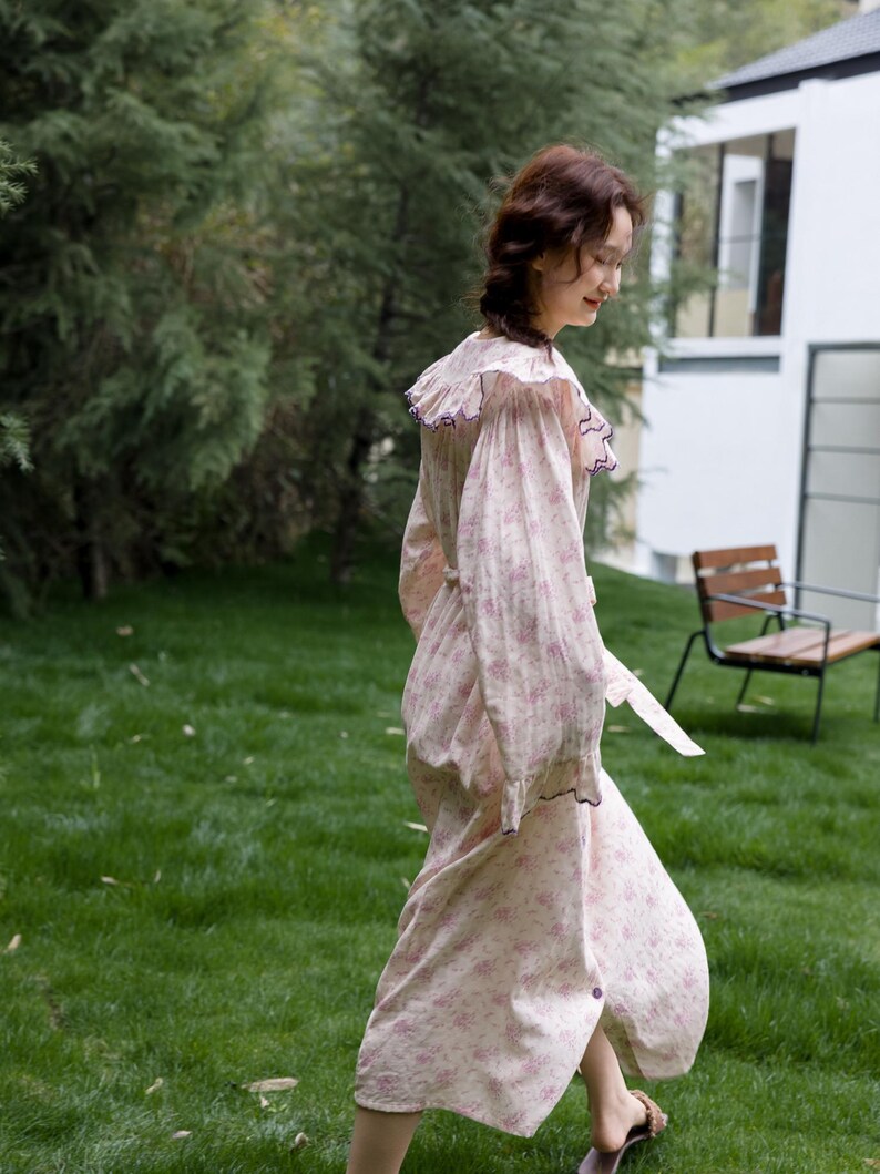 May include: A light pink floral print dress with a ruffled collar and long sleeves. The dress has a button-down front and a matching belt. The dress is worn outdoors on a grassy lawn.