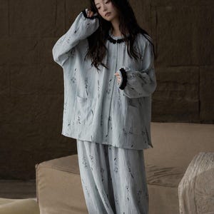 May include: Light blue pajama set with a floral pattern. The long-sleeved top has a black trim around the collar and cuffs, and two front pockets. The matching pants are loose-fitting. The fabric appears to be a soft, textured material.