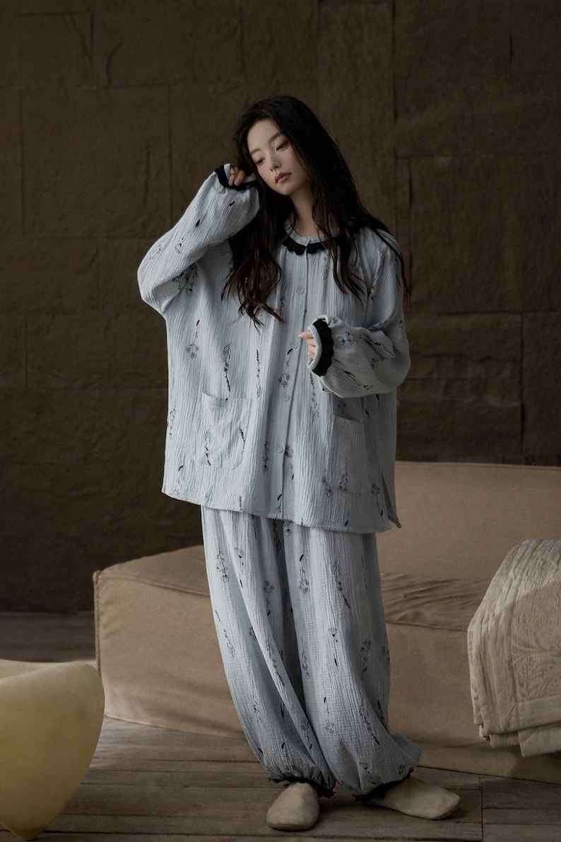 May include: Light blue pajama set with a floral pattern. The long-sleeved top has a black trim around the collar and cuffs, and two front pockets. The matching pants are loose-fitting. The fabric appears to be a soft, textured material.