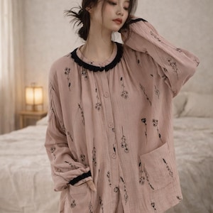 May include: A light pink pajama set with a floral pattern. The long-sleeved top has a black trim around the collar and cuffs, and a pocket on the front. The matching pants complete the set. The fabric appears soft and lightweight.