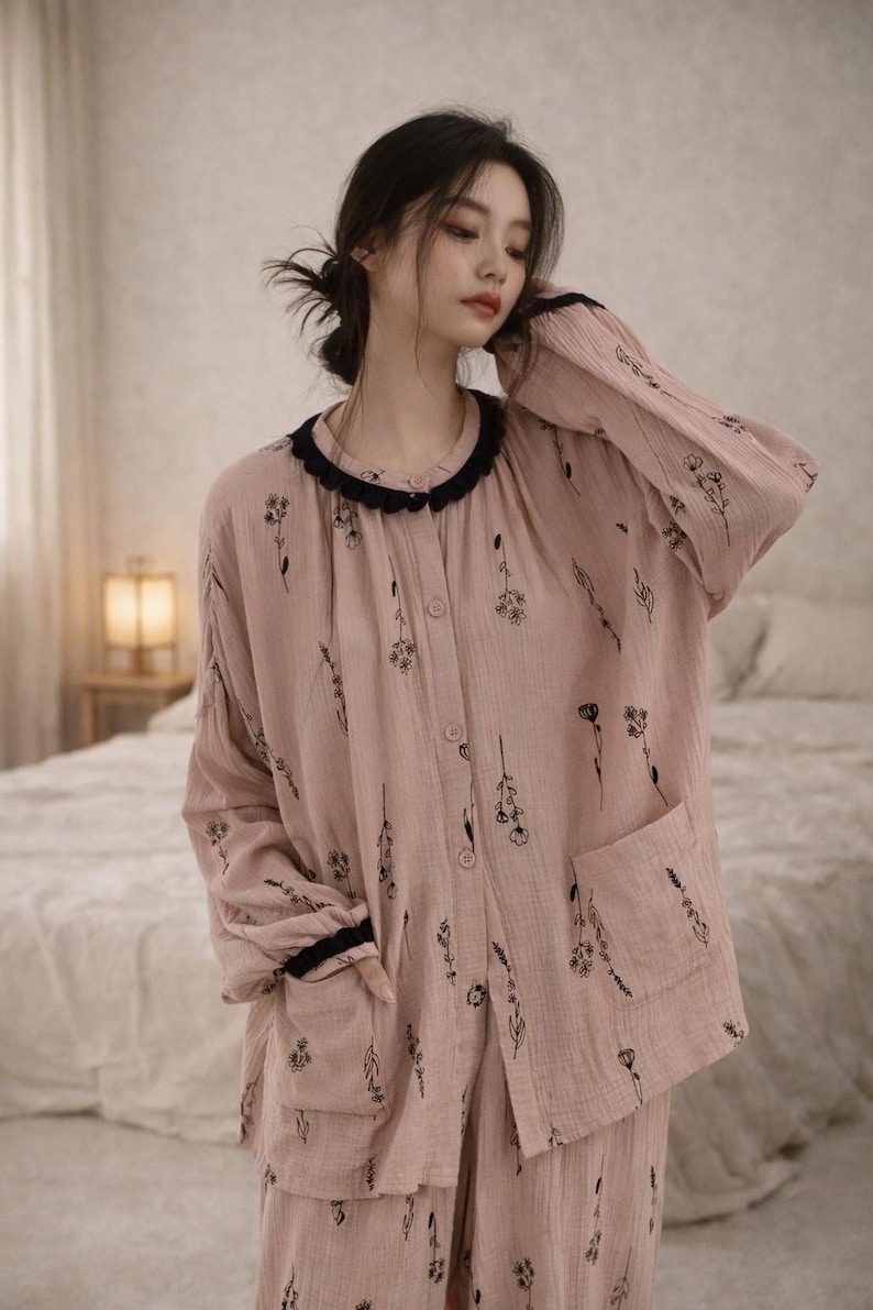 May include: A light pink pajama set with a floral pattern. The long-sleeved top has a black trim around the collar and cuffs, and a pocket on the front. The matching pants complete the set. The fabric appears soft and lightweight.