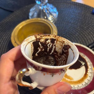 Può includere: Una piccola tazza da caffè ornata, con manico e bordo dorati, piena di fondi di caffè scuri. La tazza poggia su un piattino abbinato con un bordo bordeaux. La tazza e il piattino sono bianchi con un motivo floreale.