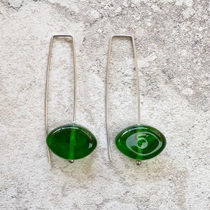 May include: A pair of dangle earrings featuring oval, deep green glass beads suspended from long, silver-toned wires. The beads have a glossy finish and a small silver bead at the bottom. The earrings are displayed against a textured, light gray background.