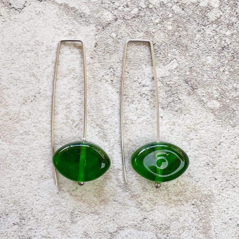 May include: A pair of dangle earrings featuring oval, deep green glass beads suspended from long, silver-toned wires. The beads have a glossy finish and a small silver bead at the bottom. The earrings are displayed against a textured, light gray background.