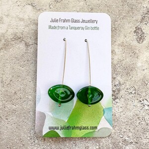 May include: A pair of green glass earrings with long silver hooks. The earrings are oval-shaped and have a glossy finish. The card behind the earrings reads "Julie Frahm Glass Jewellery Made from a Tanqueray Gin bottle" and "www.juliefrahmglass.com".