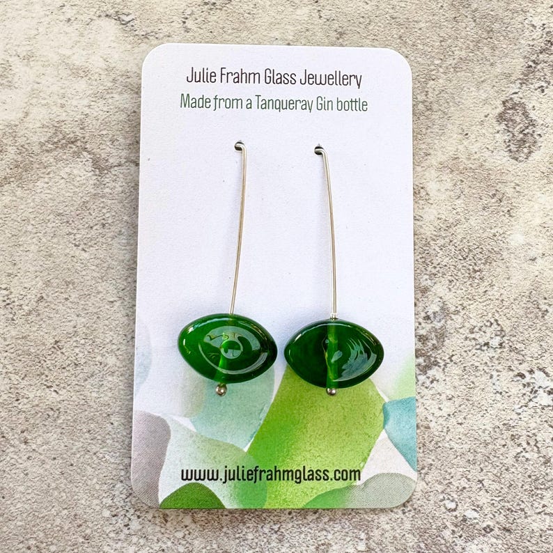 May include: A pair of green glass earrings with long silver hooks. The earrings are oval-shaped and have a glossy finish. The card behind the earrings reads "Julie Frahm Glass Jewellery Made from a Tanqueray Gin bottle" and "www.juliefrahmglass.com".