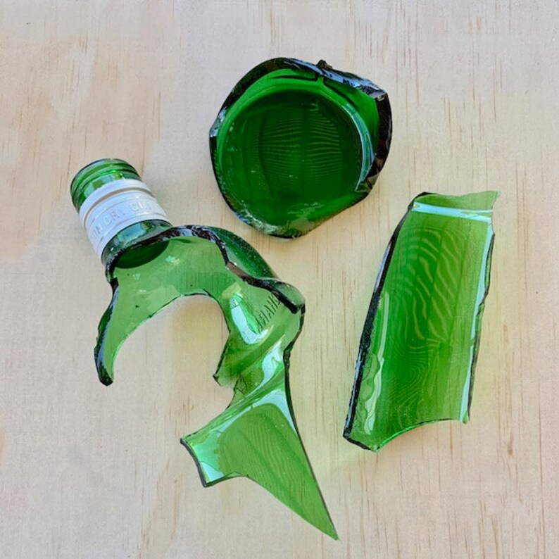 May include: Shattered green glass bottle fragments, including the neck with a white label and the intact green cap. The broken pieces are scattered on a light wood surface, showcasing the bottle's original form.
