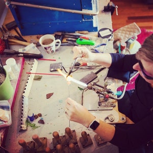 May include: A person working with glass, using a torch to heat and shape it. The workspace includes tools, a blue machine, and a coffee mug. The person is wearing safety glasses and a black shirt. The image is taken from an overhead perspective.