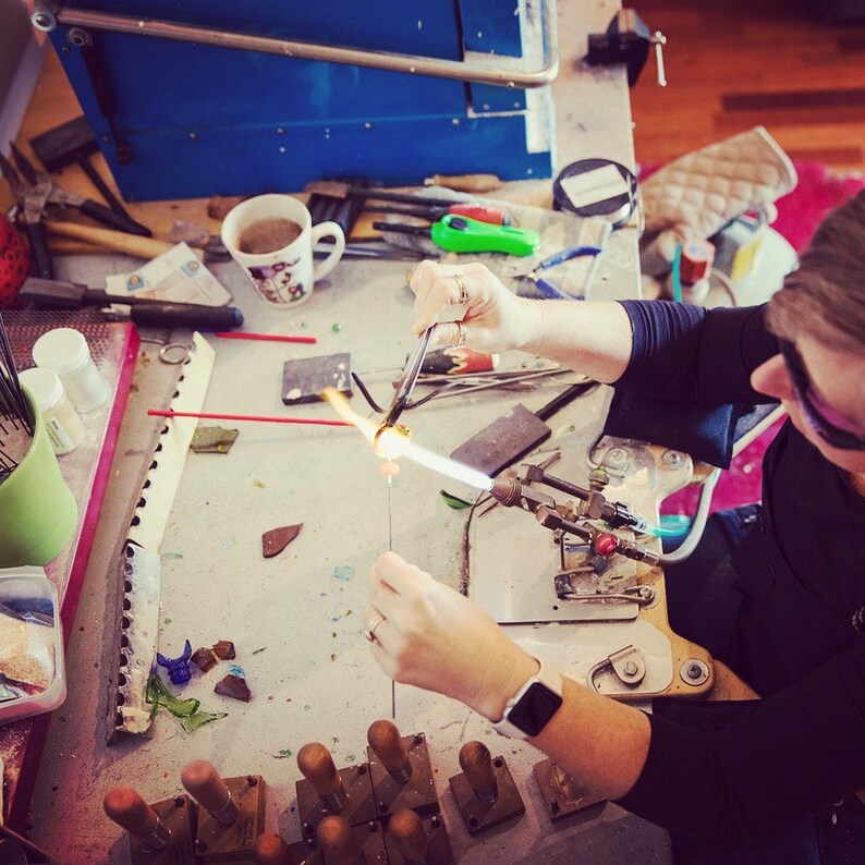 May include: A person working with glass, using a torch to heat and shape it. The workspace includes tools, a blue machine, and a coffee mug. The person is wearing safety glasses and a black shirt. The image is taken from an overhead perspective.