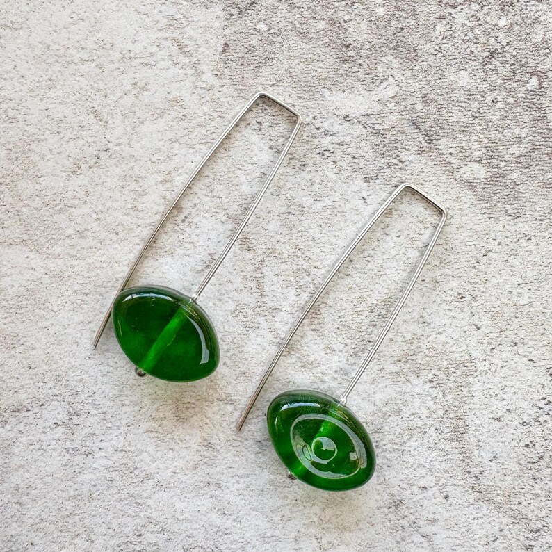 May include: A pair of dangle earrings featuring oval-shaped, deep green glass beads. The beads are suspended from long, silver-colored, rectangular wire hooks. The earrings are displayed on a light gray, textured surface.