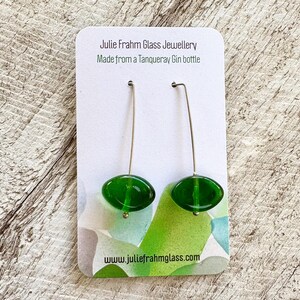May include: A pair of handmade green glass earrings with silver-colored ear wires. The earrings are oval-shaped and suspended from a card that reads "Julie Frahm Glass Jewellery Made from a Tanqueray Gin bottle" and "www.juliefrahmglass.com".