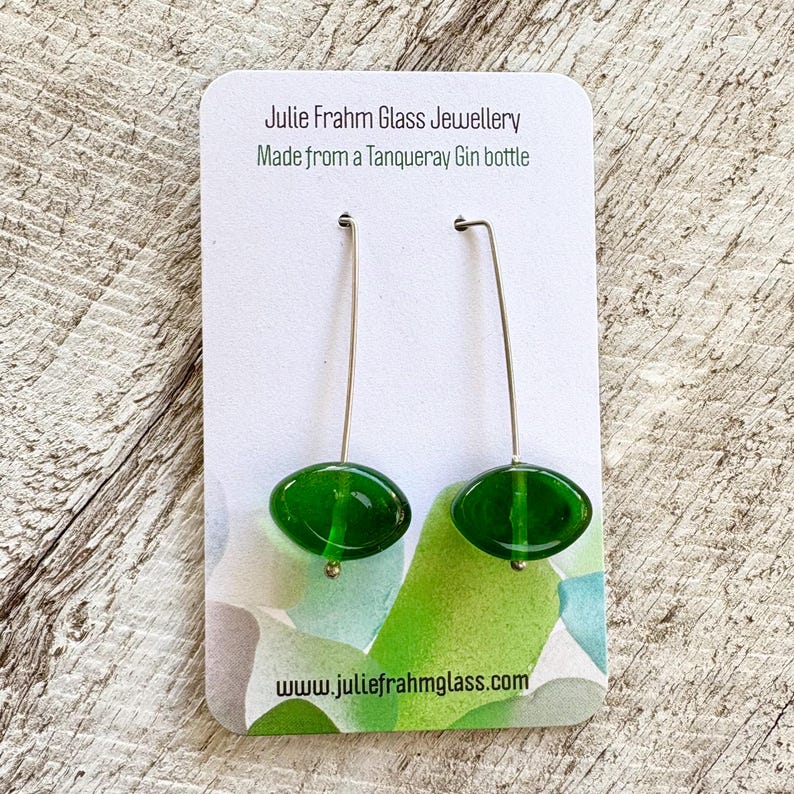 May include: A pair of handmade green glass earrings with silver-colored ear wires. The earrings are oval-shaped and suspended from a card that reads "Julie Frahm Glass Jewellery Made from a Tanqueray Gin bottle" and "www.juliefrahmglass.com".