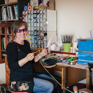 May include: A woman in a workshop smiles while holding a glassblowing tool. A variety of colorful glass pieces are displayed on a rack behind her. The workspace includes tools, a wooden desk, and a blue toolbox.