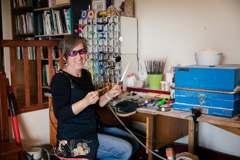 May include: A woman in a workshop smiles while holding a glassblowing tool. A variety of colorful glass pieces are displayed on a rack behind her. The workspace includes tools, a wooden desk, and a blue toolbox.