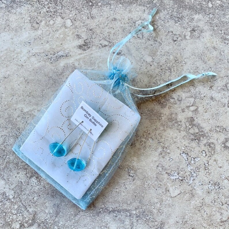 May include: A pair of handmade earrings with light blue glass beads, presented in a sheer blue gift bag. The earrings are on a white card that reads "Bombay Sapphire Gin Bottle."