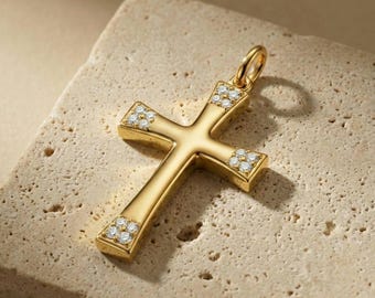 May include: Gold-colored cross pendant with small, clear gemstones set at the ends of each arm. The pendant has a loop at the top for a chain. The cross is displayed on a textured, light-colored surface.