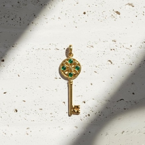 Op de afbeelding: Een goudkleurige sleutelhanger met een cirkelvormige bovenkant met een bloemmotief met groene edelstenen. De sleutel heeft een lange steel en een gedetailleerd sleutelgatontwerp aan de onderkant. De hanger wordt weergegeven tegen een getextureerde, gebroken witte achtergrond.