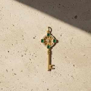 Op de afbeelding: Een goudkleurige sleutelhanger met een decoratieve kop met een bloemmotief, smaragdgroene edelstenen en een centrale diamant. De schacht en baard van de sleutel zijn ook goudkleurig.