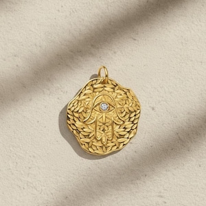 Op de afbeelding: Een goudkleurige hanger met een Hamsa-handontwerp, met een oog met een kleine, heldere edelsteen in het midden. De hanger is omgeven door een bladerpatroon en heeft een lus aan de bovenkant voor een ketting.