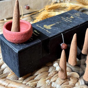 May include: A red ceramic incense holder with a brown incense cone, next to a black box of cone incense. The box has gold text that reads "Handmade in Nepal". Several brown incense cones are visible.