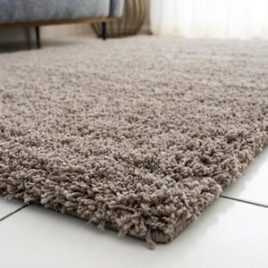 May include: A rectangular area rug with a thick, high-pile shag texture in a solid light taupe or beige color. The rug features long, soft fibers that create a dense, plush surface, covering a portion of a smooth, white tiled floor surface.