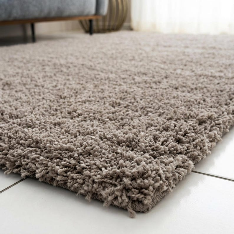 May include: A rectangular area rug with a thick, high-pile shag texture in a solid light taupe or beige color. The rug features long, soft fibers that create a dense, plush surface, covering a portion of a smooth, white tiled floor surface.