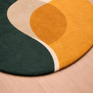 May include: A round rug with an abstract design. The rug features a dark green, tan, and mustard yellow color palette. The design includes curved shapes and a white border. The rug is on a wooden floor.