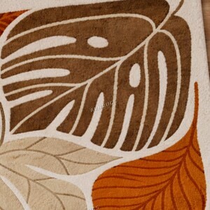May include: A close-up of a beige rug with a leaf design in shades of brown and orange. The rug features a large, stylized leaf in the centre, with smaller leaves in the foreground. The text "APNRUGS" is visible on the rug.