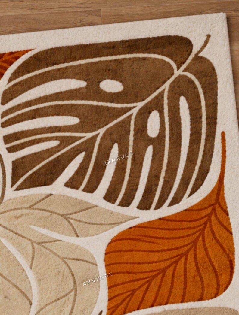 May include: A close-up of a beige rug with a leaf design in shades of brown and orange. The rug features a large, stylized leaf in the centre, with smaller leaves in the foreground. The text "APNRUGS" is visible on the rug.