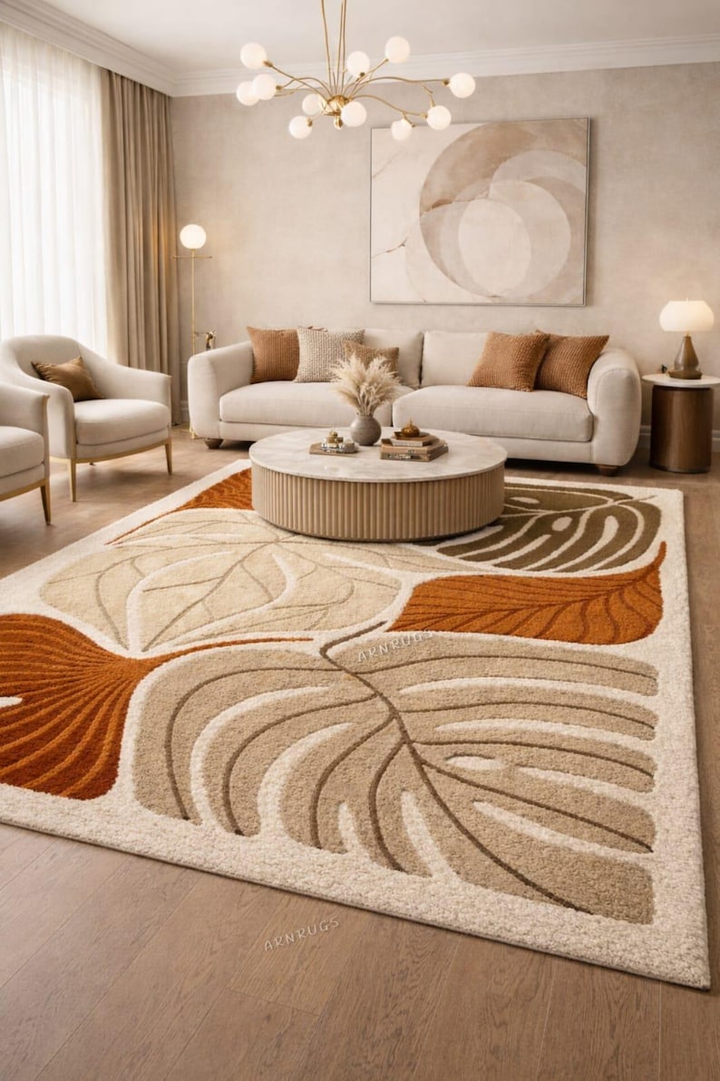 May include: A large rug featuring a leaf pattern in beige, brown, and orange tones. The rug is in a living room with a cream-colored sofa, armchairs, and a circular coffee table. The room has a neutral colour scheme.