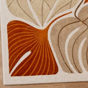 May include: Close-up of a beige and orange patterned rug. The rug features a stylized leaf design with curved lines and a white border. The rug has the text "ARN RUGS" in the lower right corner.