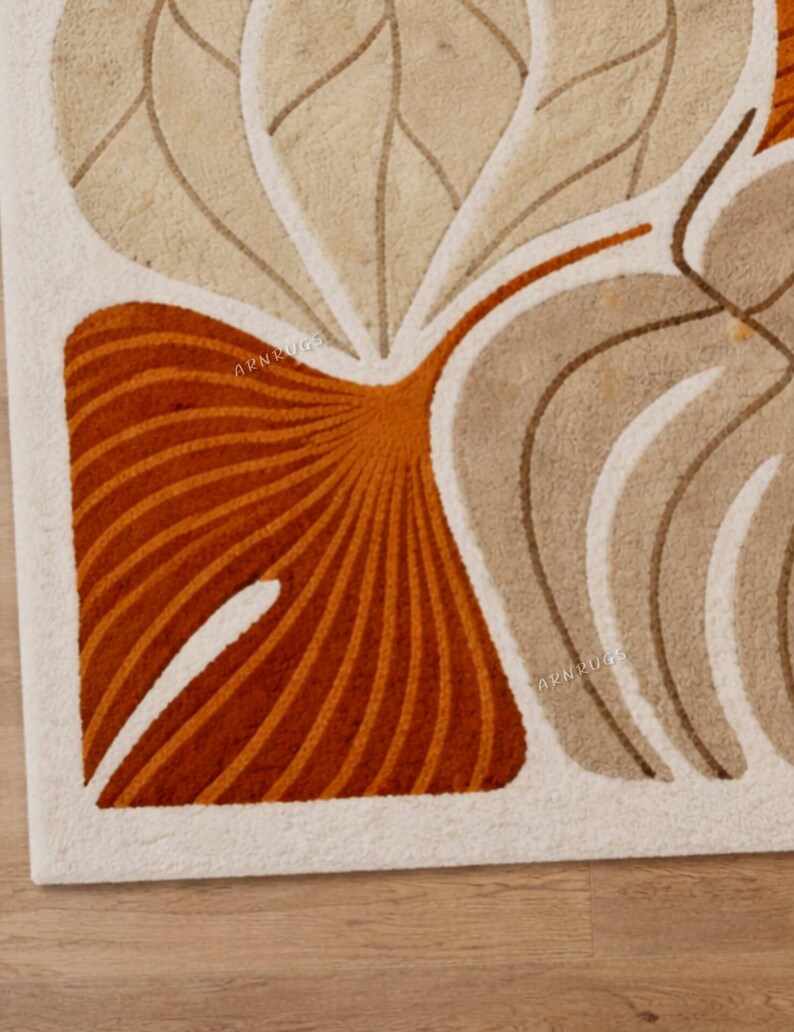 May include: Close-up of a beige and orange patterned rug. The rug features a stylized leaf design with curved lines and a white border. The rug has the text "ARN RUGS" in the lower right corner.