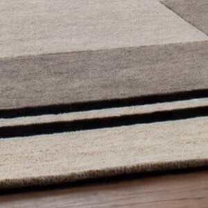 May include: A rectangular area rug with a geometric design. The rug features a combination of beige, gray, and brown tones, with two bold black stripes. The rug is made of a textured material, likely wool or a similar fabric.