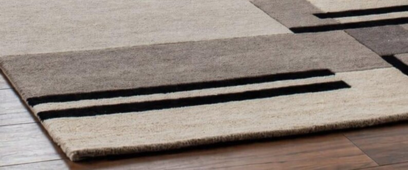 May include: A rectangular area rug with a geometric design. The rug features a combination of beige, gray, and brown tones, with two bold black stripes. The rug is made of a textured material, likely wool or a similar fabric.