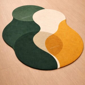 May include: A modern, abstract area rug with a unique, organic shape. The rug features overlapping sections of dark green, beige, cream, and mustard yellow. The text "ARNRUGS" is visible on the rug.
