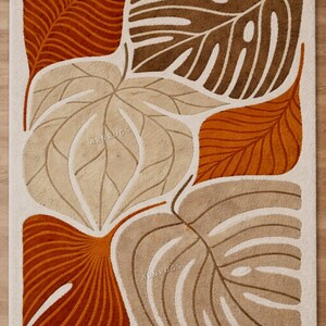 May include: A rectangular rug featuring a cream backdrop and a design of oversized, stylised leaves in brown, tan, and orange hues. The leaves display intricate vein patterns, and the overall aesthetic evokes mid-century modern art.