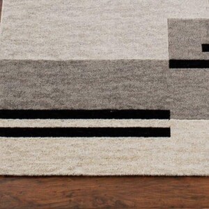 May include: A rectangular area rug with a geometric design. The rug features a combination of light beige, gray, and black rectangular shapes. Two black stripes run horizontally across the bottom of the rug.
