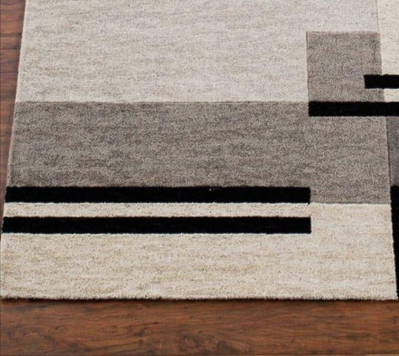May include: A rectangular area rug with a geometric design. The rug features a combination of light beige, gray, and black rectangular shapes. Two black stripes run horizontally across the bottom of the rug.