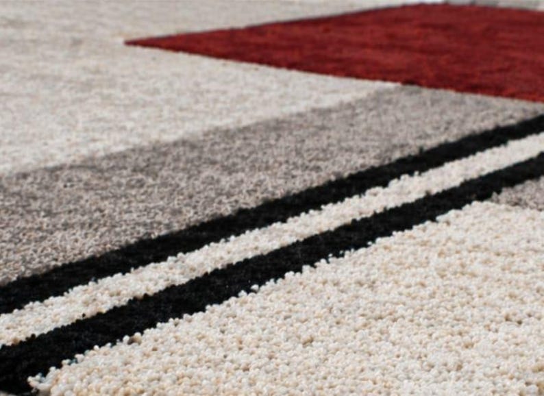 May include: Close-up of a geometric patterned rug. The rug features a cream, gray, and burgundy color scheme with black stripes. The design includes a burgundy triangle and two black parallel lines.