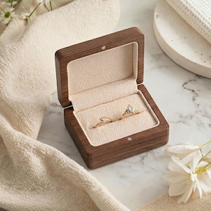 May include: A wooden ring box with a light beige interior, holding two gold rings. One ring features a prominent diamond. The box is open, revealing the rings. The scene is set on a marble surface with flowers and a folded cloth.