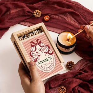 Könnte beinhalten: Eine rechteckige Streichholzschachtel aus Holz mit Schiebedeckel, verziert mit einem rosa gestreiften Etikett, einer roten Schleife, Weinglas-Illustrationen und dem Text ANNIE'S 30th BIRTHDAY. Die Schachtel enthält Streichhölzer mit dunklen Köpfen.